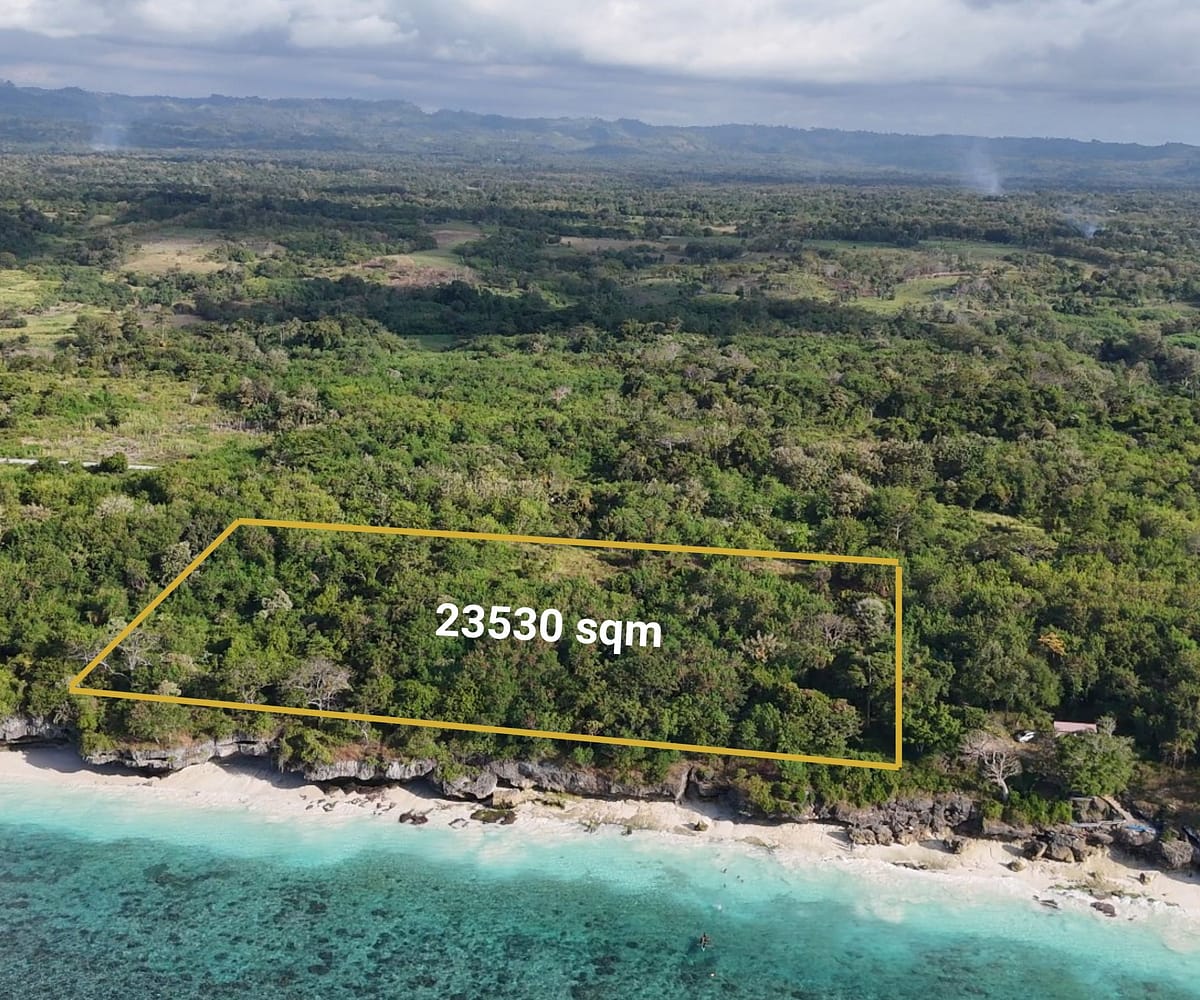 A spectacular high-angle aerial view showcasing the property's dramatic, elevated rocky crest dropping directly down onto a pristine, untouched white sand beach meeting clear turquoise coral waters.