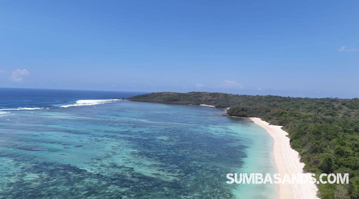 An expansive high-angle view showcasing the striking color contrast of the calm bay. The crystal-clear, shallow turquoise waters reveal beautifully textured coral reefs below the surface, stretching out toward the deep blue horizon under a clear sky.