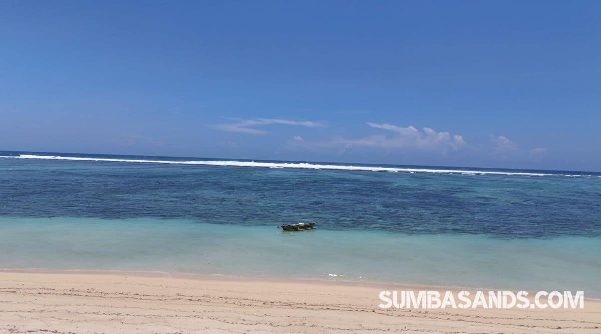 An expansive high-angle view showcasing the striking color contrast of the calm bay. The crystal-clear, shallow turquoise waters reveal beautifully textured coral reefs below the surface, stretching out toward the deep blue horizon under a clear sky.