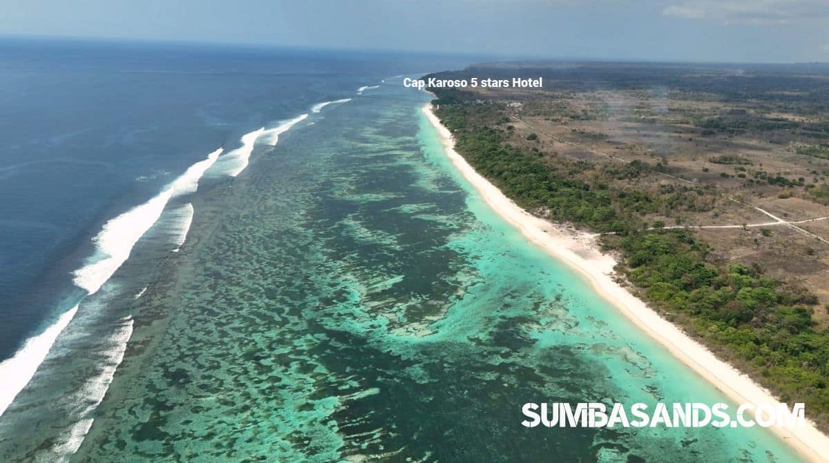 A breathtaking aerial drone photograph overlooking the pristine coastline of Kodi in West Sumba. The image captures vibrant turquoise ocean waters and shallow coral reefs gently meeting a bright white sandy beach, bordered by lush tropical green vegetation.