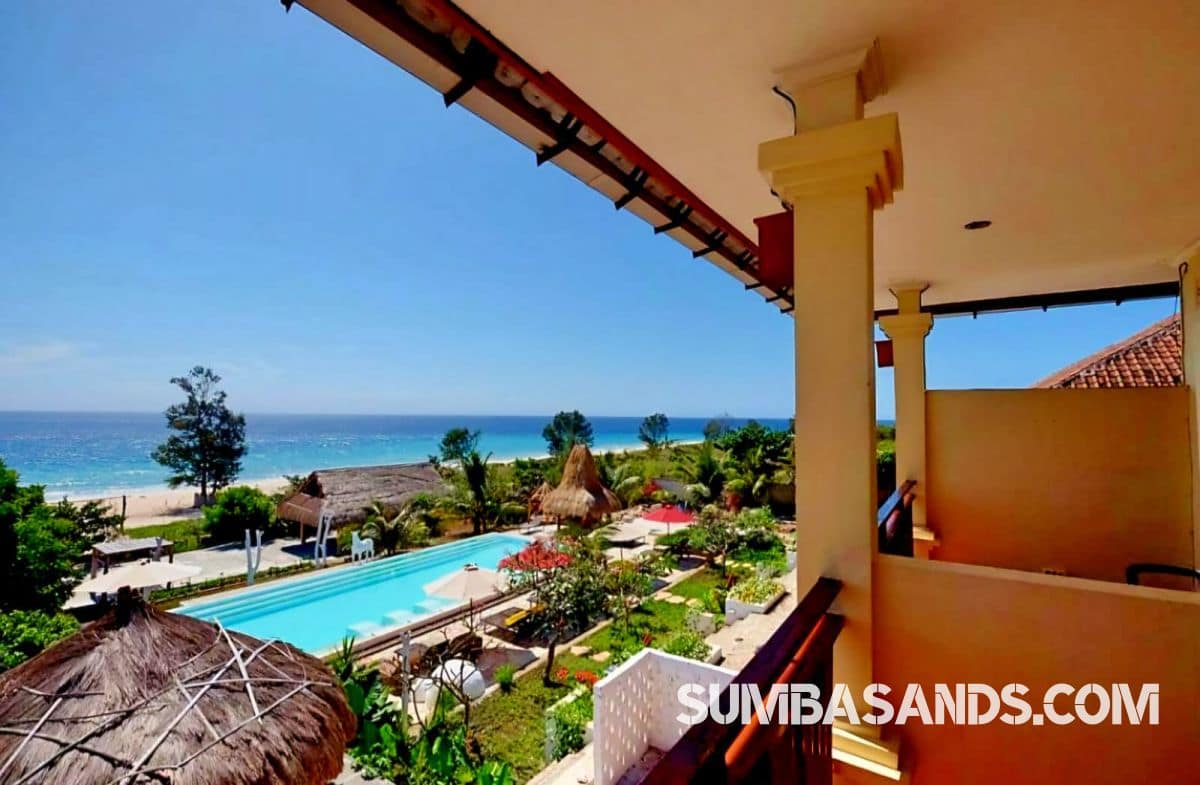 : An aerial drone photograph capturing the complete beachfront layout of this turnkey estate in West Sumba. The image showcases a pristine 100-meter white sand beach leading up to a resort footprint complete with an emerald swimming pool, tropical landscaping, and a large central building structure.
