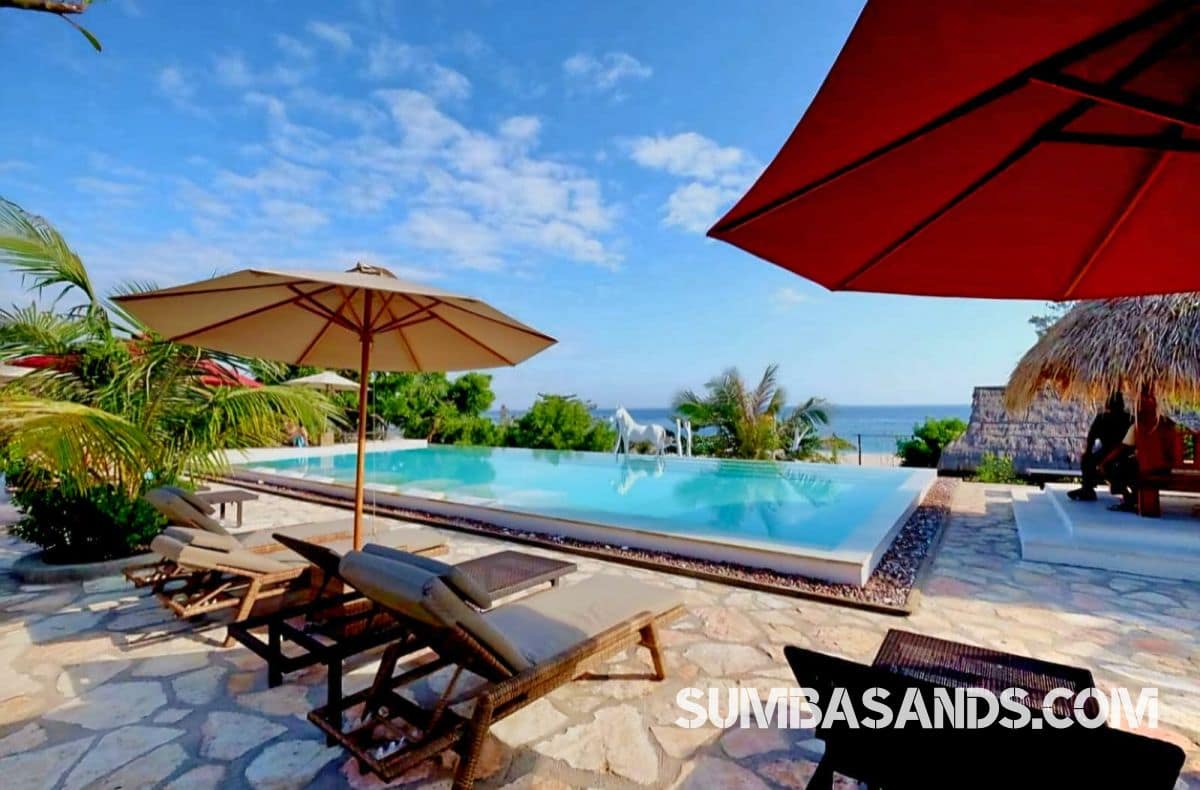 : An aerial drone photograph capturing the complete beachfront layout of this turnkey estate in West Sumba. The image showcases a pristine 100-meter white sand beach leading up to a resort footprint complete with an emerald swimming pool, tropical landscaping, and a large central building structure.