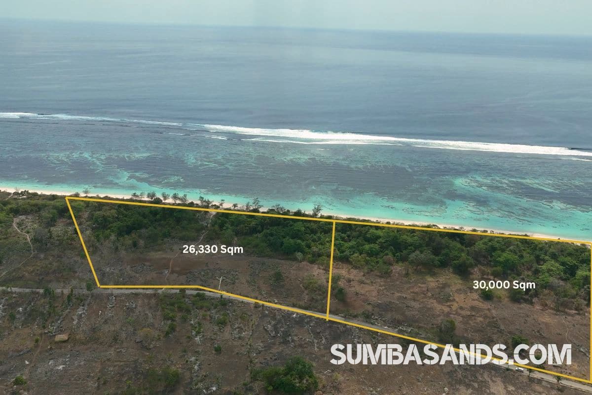 A breathtaking aerial drone photograph overlooking the pristine coastline of Kodi in West Sumba. The image captures vibrant turquoise ocean waters and shallow coral reefs gently meeting a bright white sandy beach, bordered by lush tropical green vegetation.