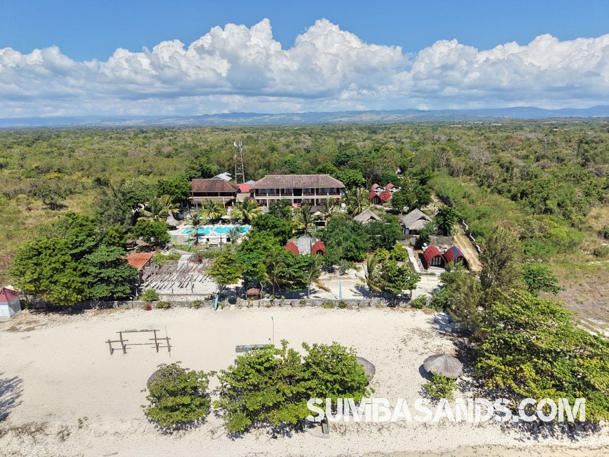 : An aerial drone photograph capturing the complete beachfront layout of this turnkey estate in West Sumba. The image showcases a pristine 100-meter white sand beach leading up to a resort footprint complete with an emerald swimming pool, tropical landscaping, and a large central building structure.