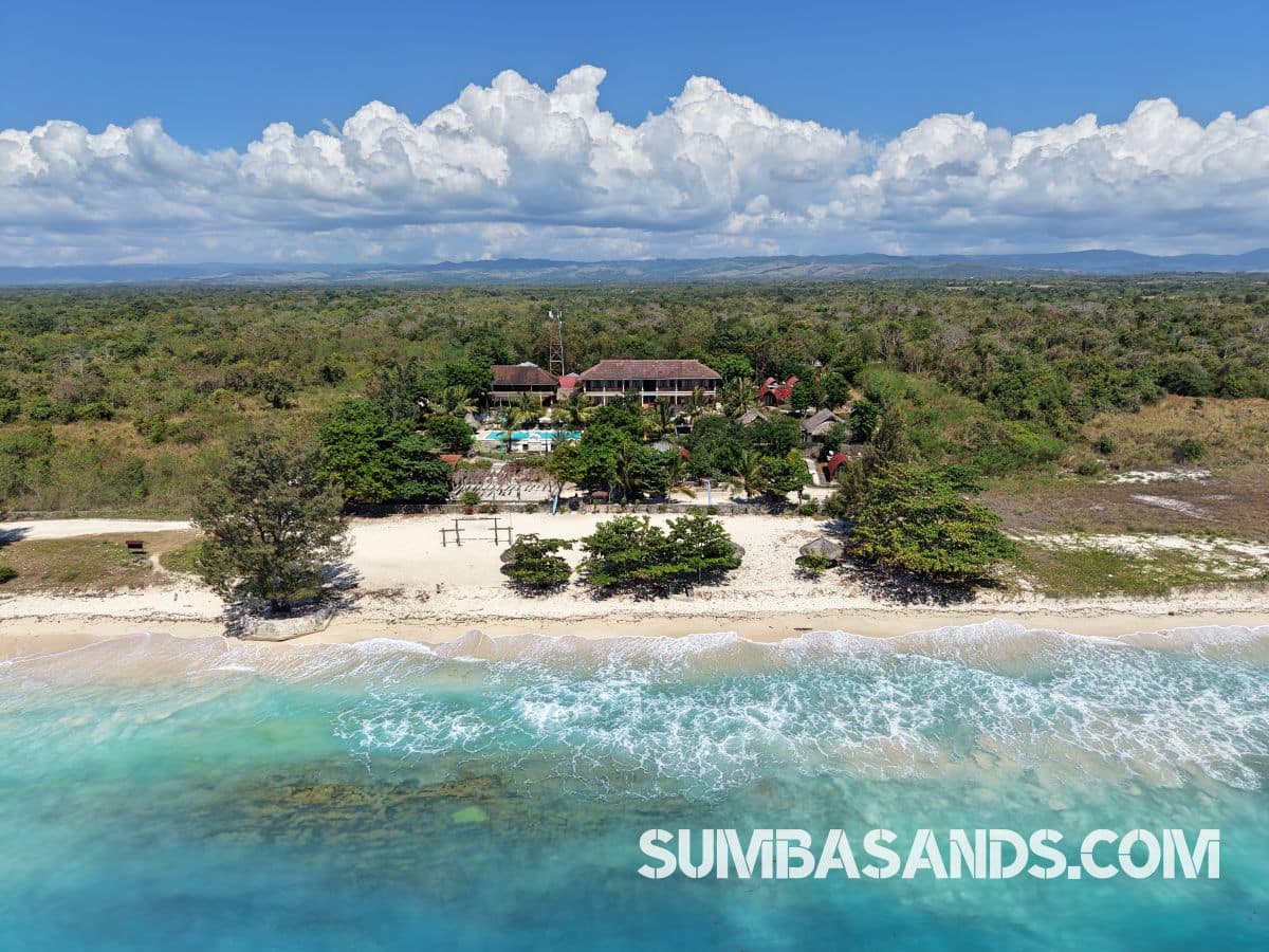 : An aerial drone photograph capturing the complete beachfront layout of this turnkey estate in West Sumba. The image showcases a pristine 100-meter white sand beach leading up to a resort footprint complete with an emerald swimming pool, tropical landscaping, and a large central building structure.