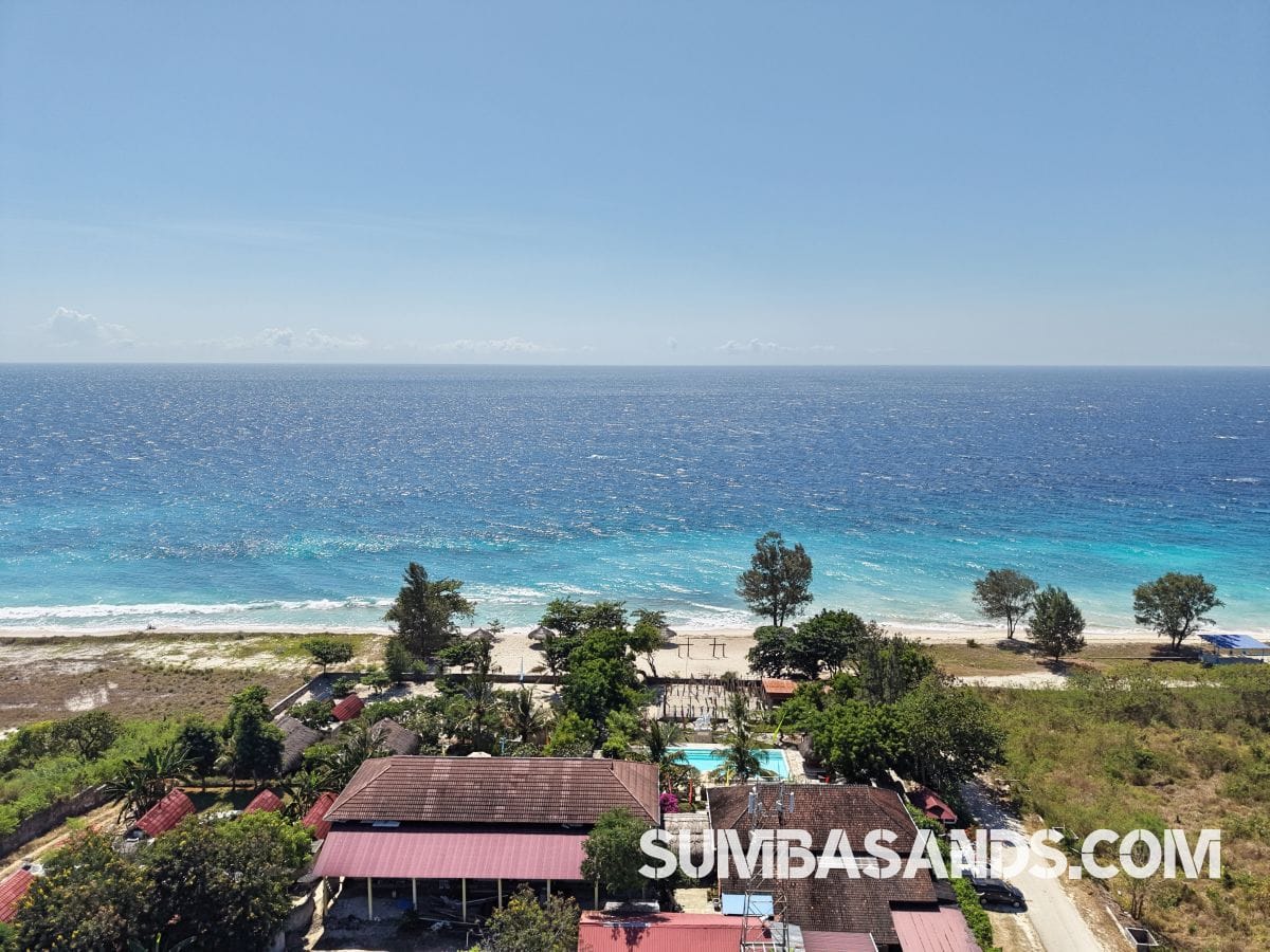: An aerial drone photograph capturing the complete beachfront layout of this turnkey estate in West Sumba. The image showcases a pristine 100-meter white sand beach leading up to a resort footprint complete with an emerald swimming pool, tropical landscaping, and a large central building structure.