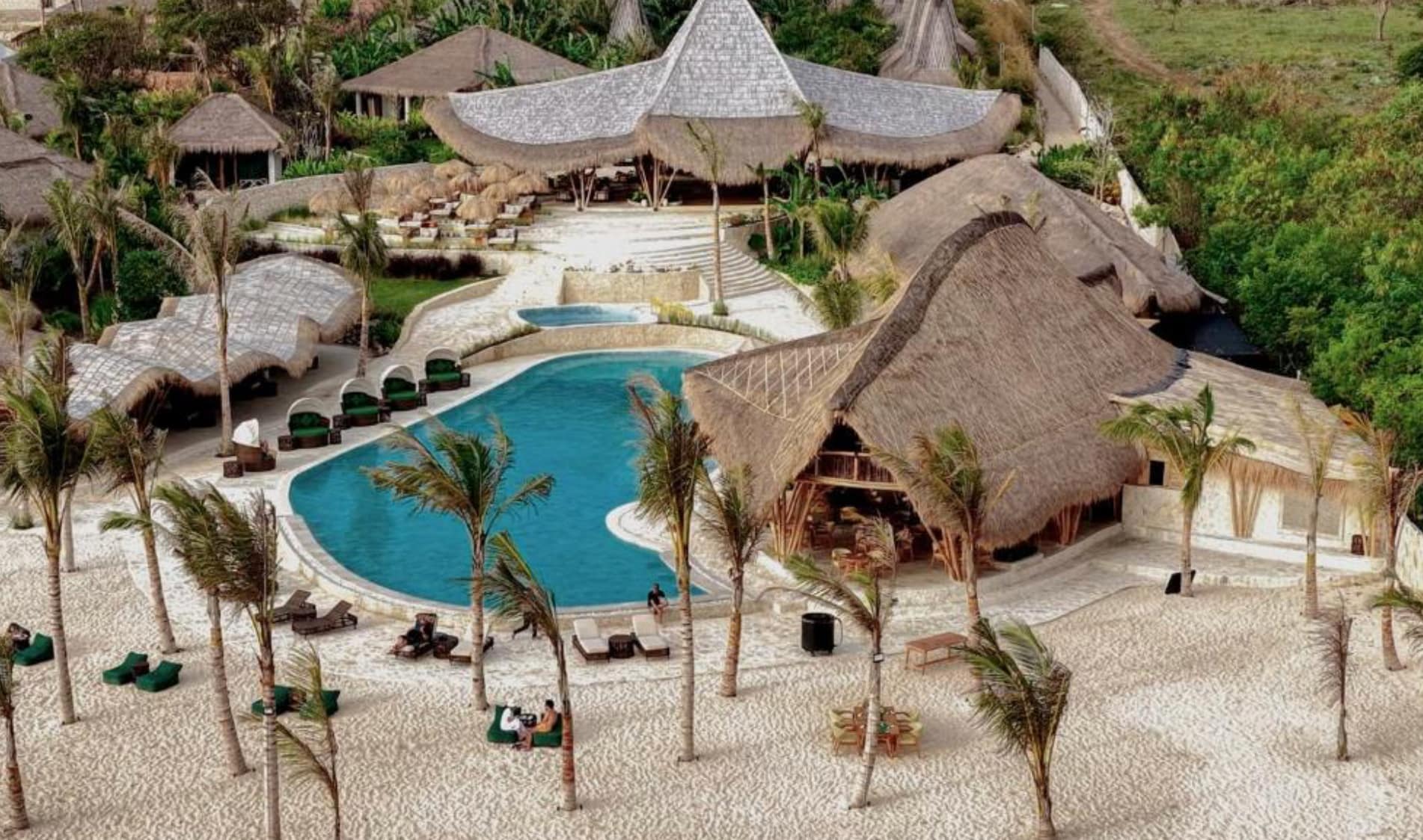 An aerial perspective of the Arya Sumba resort in Indonesia, showing traditional-style thatched-roof villas clustered along a sandy beach next to a turquoise ocean, surrounded by dense tropical foliage.