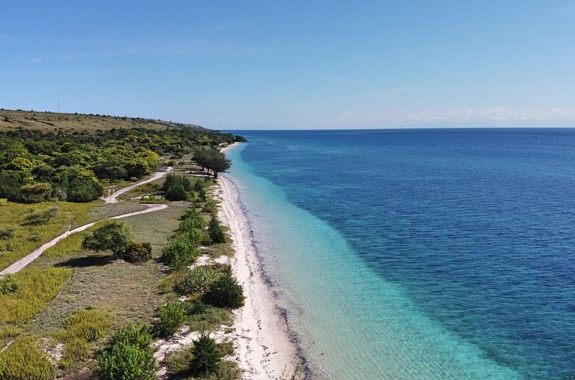 A wide aerial shot of the 2-hectare Hamba Praing Beachfront Estate. The image captures the flat, pristine land meeting the turquoise waters of the Savu Sea, with the golden savanna hills of Hamba Praing in the background