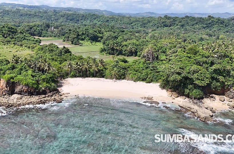 A panoramic aerial view of the Beralewa Secluded Beach Estate. The image captures 1.7 hectares of lush green land meeting a wide, deep white-sand beach with turquoise waters, located along the Southwest Sumba coastline