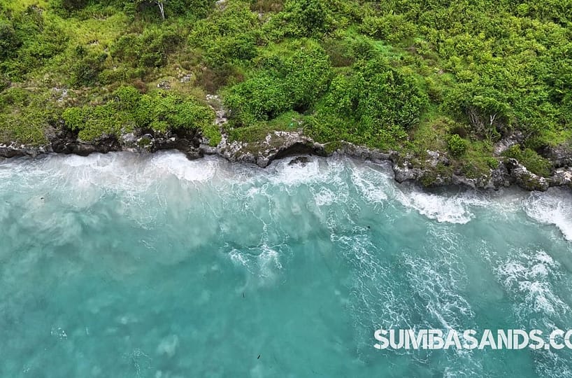 A panoramic aerial view of the Kawona Horizon Development Plot. The image captures elevated green terrain overlooking the white sands of Kawona Beach, with the clear blue waters of the Savu Sea and a paved asphalt road bordering the property.